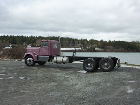 1959 Kenworth W900A - Semi-truck chassis with a purple cab sits by the water under a cloudy sky.