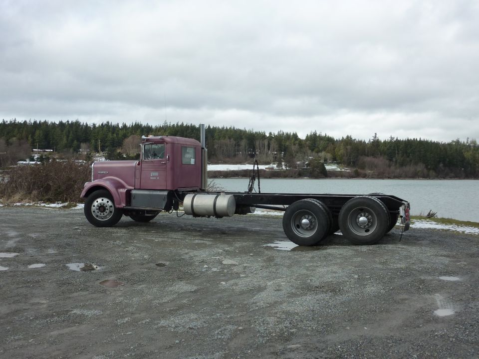 Purple semi-truck chassis parked on gravel by a body of water, with trees in the background