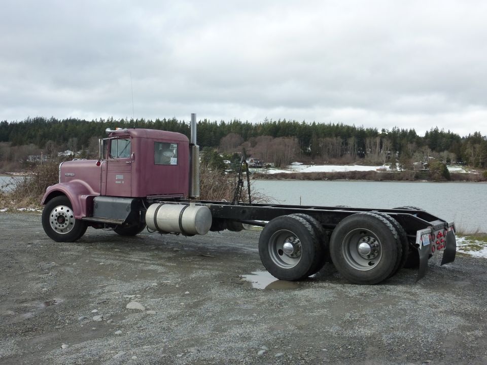 Purple semi-truck chassis parked by a body of water. It has a single cab and a long frame with dual rear wheels