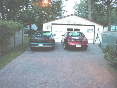 1997 Ford Taurus - Two sedans are parked in a driveway in front of a garage, one dark green and one red.