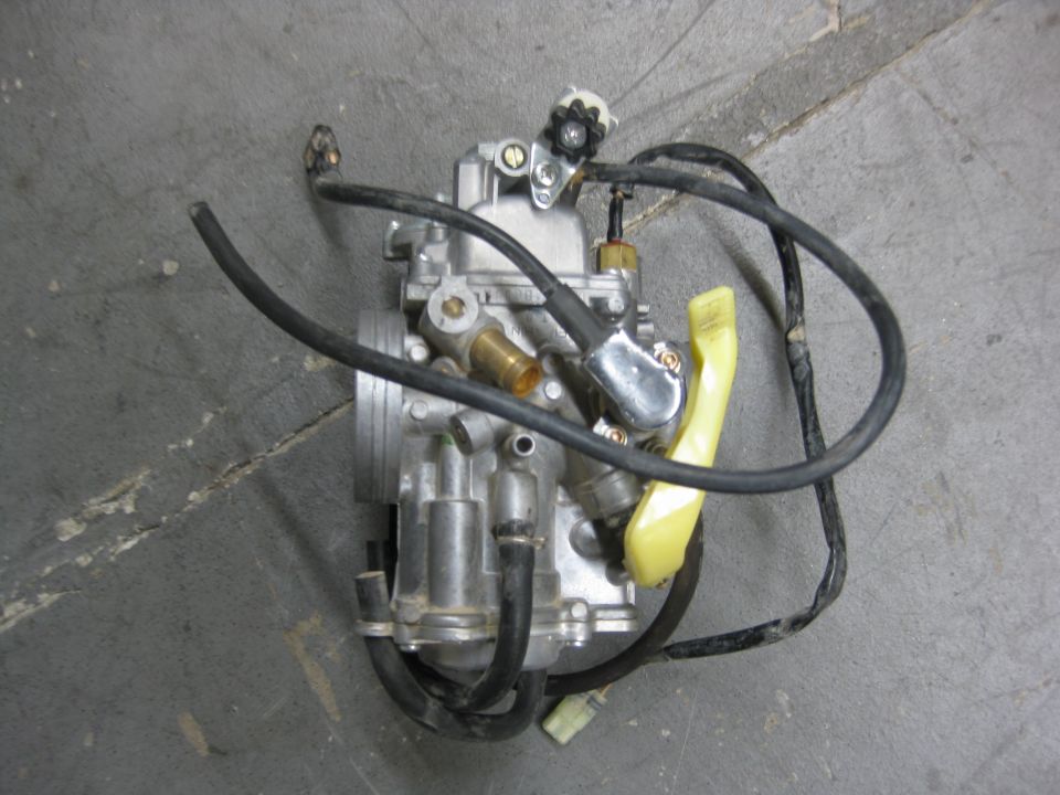 Removed aluminum carburetor with black hoses and yellow lever lying on a concrete floor