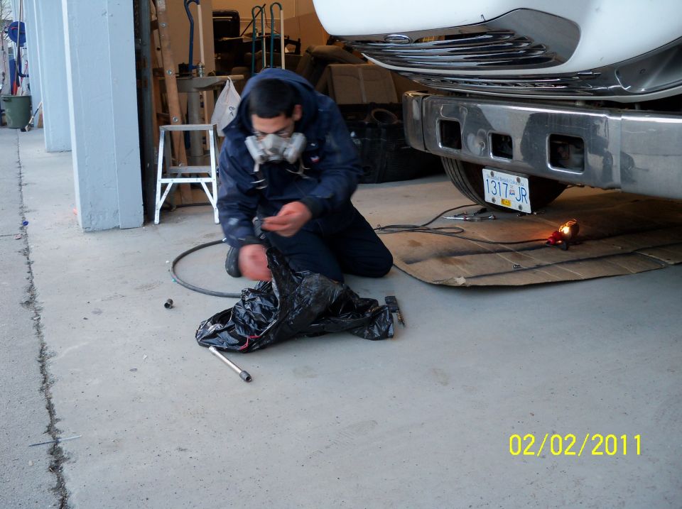 Man in respirator works on vehicle rear bumper and undercarriage. License plate visible