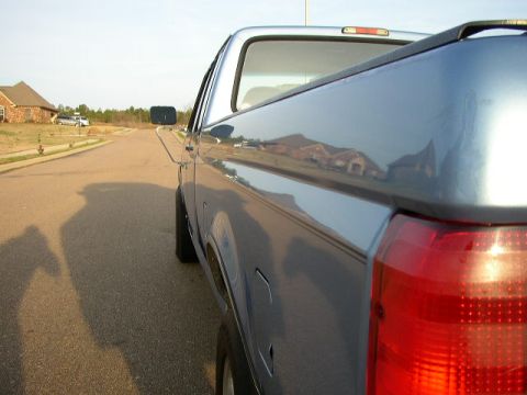 1995 Ford Bronco - Side view of a blue pickup truck, with reflections of houses and trees visible on its body panels.