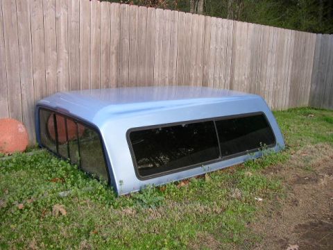 1995 Ford Bronco - A truck bed cap rests on the grass next to a weathered wooden fence.