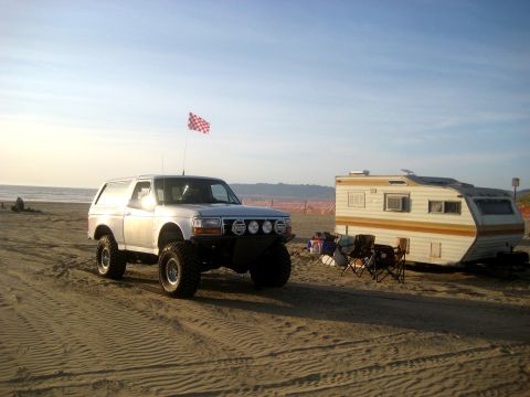 1996 Ford Bronco - A modified 1996 Ford Bronco sits on a sandy beach next to a trailer, with a checkered flag flying from its antenna.