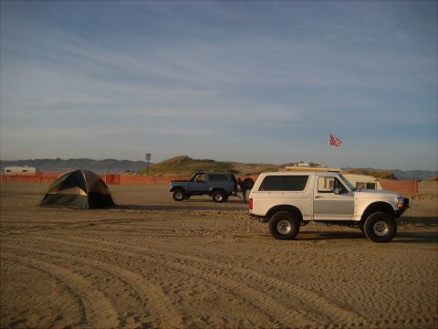 1996 Ford Bronco - Two Ford Broncos and a tent are on a sandy beach under a partly cloudy sky.