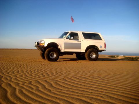 1996 Ford Bronco - A white 1996 Ford Bronco is parked on rippled sand dunes under a clear blue sky, with the ocean visible in the distance.