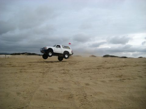 1996 Ford Bronco - A white 1996 Ford Bronco is captured mid-air while jumping over sand dunes under a cloudy sky.