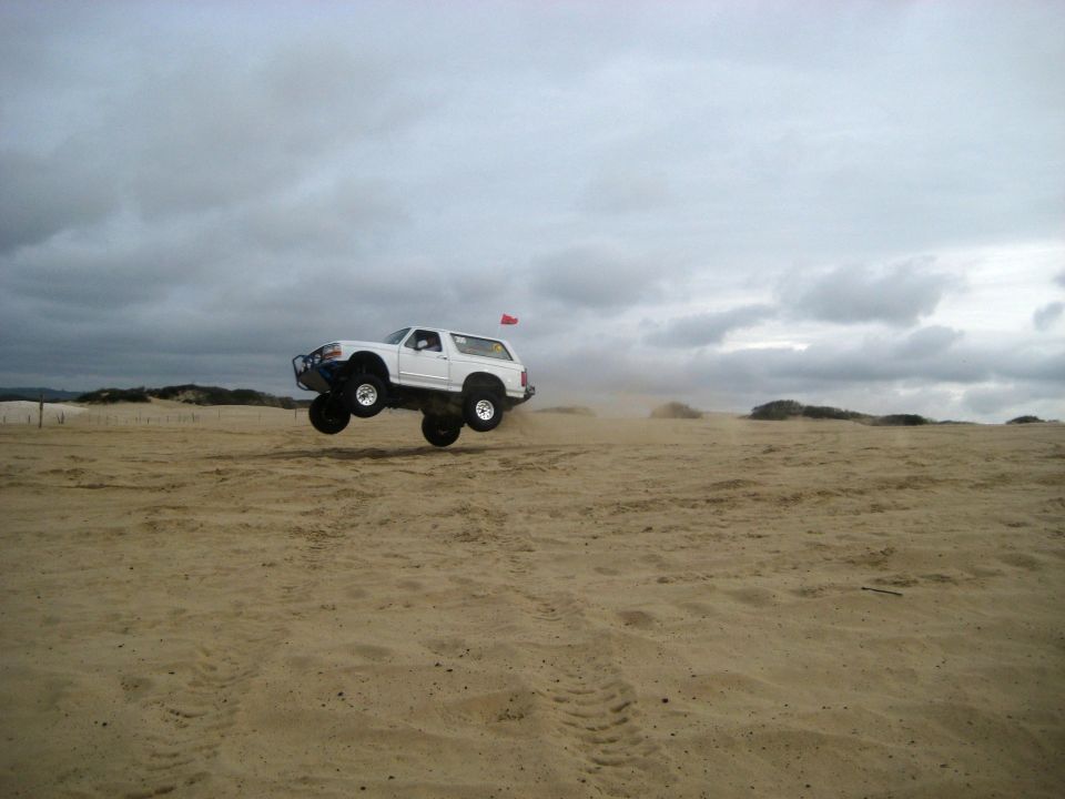 White 1996 Ford Bronco airborne over sand dunes with a red flag on top