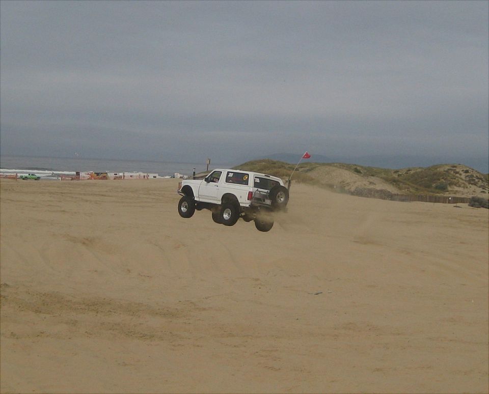 White 1992 Ford Bronco jumping over a sand dune, rear-mounted spare tire visible