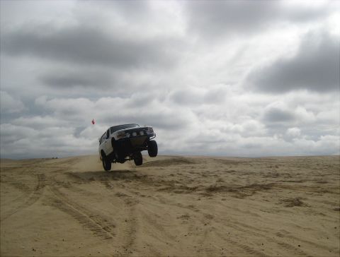 1996 Ford Bronco - A white 1996 Ford Bronco catches air while driving over sand dunes on a cloudy day.