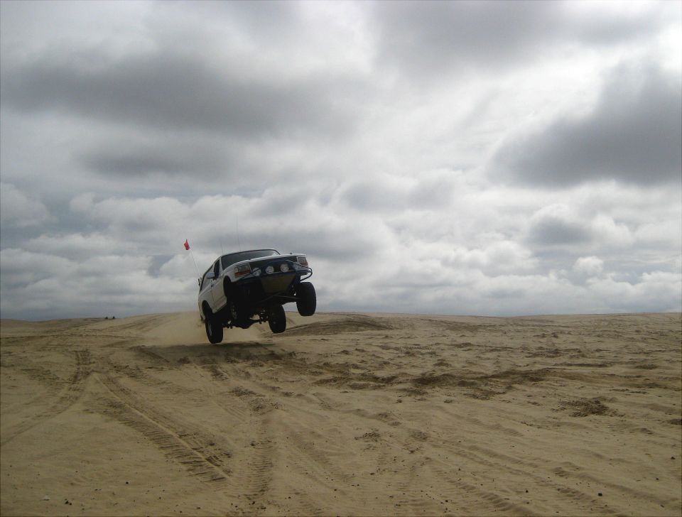 White 1996 Ford Bronco airborne over sand dunes under cloudy sky
