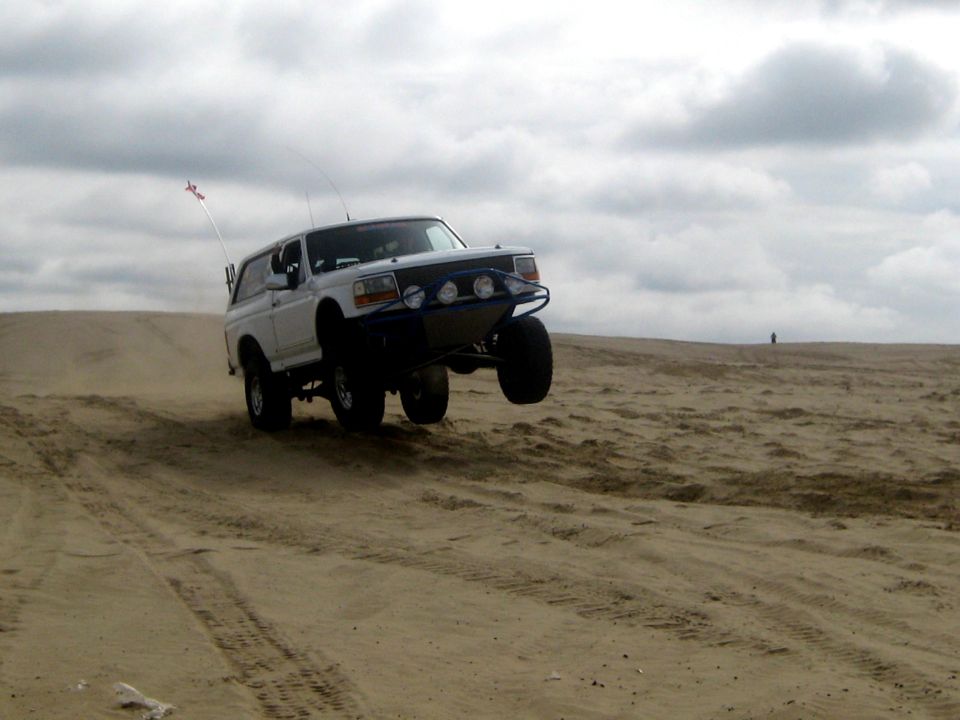 White 1996 Ford Bronco airborne over sand dune with aftermarket bumper and lights