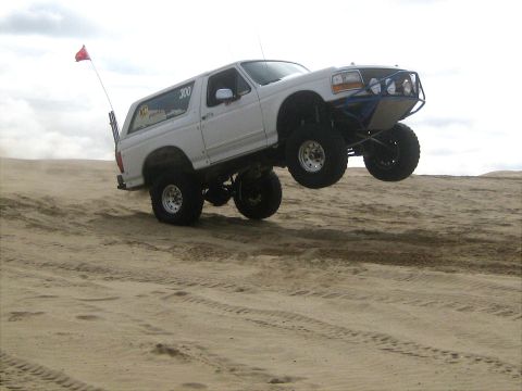 1996 Ford Bronco - White Ford Bronco with custom blue roll cage and large tires airborne over sand dunes.