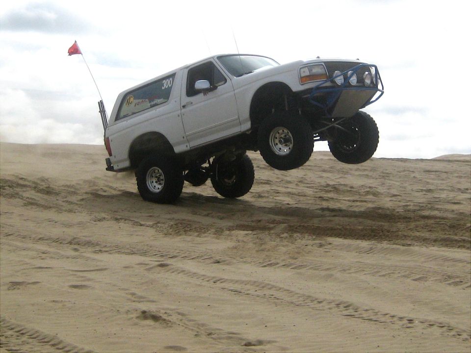White Ford Bronco with custom blue roll cage and large tires airborne over sand dunes