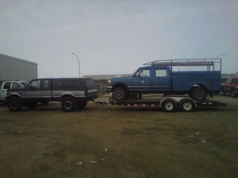 1994 Ford F-150 - A lifted pickup truck tows a blue crew cab pickup truck on a trailer under an overcast sky.