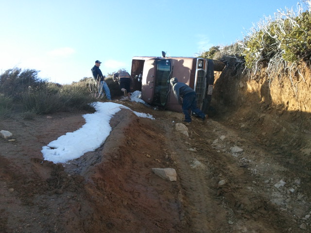 Brown 1978 Ford Bronco on its side on a dirt track with snow patches, people around it