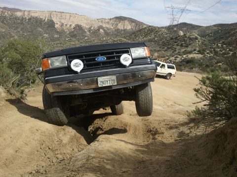 1978 Ford Bronco - A black Ford Bronco tackles a challenging off-road path, with another white SUV visible in the background.