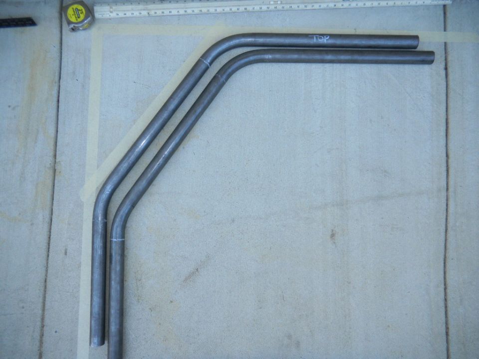 three bent metal tubes laid out on a concrete surface, one has 'TOP' written on it