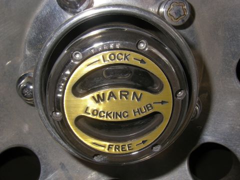 1990 Ford Bronco - Close-up of a Warn manual locking hub installed in a polished aluminum wheel center, with a gold anodized dial reading "WARN LOCKING HUB" a…