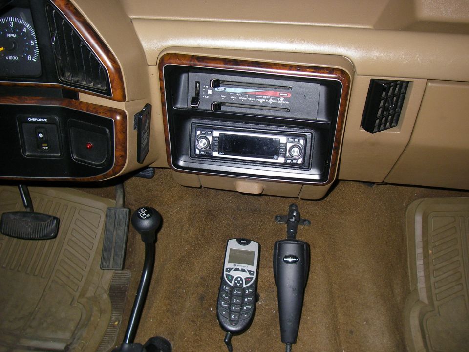 Tan vehicle interior with aftermarket stereo, wood trim, shifter, phone and mic on carpet.