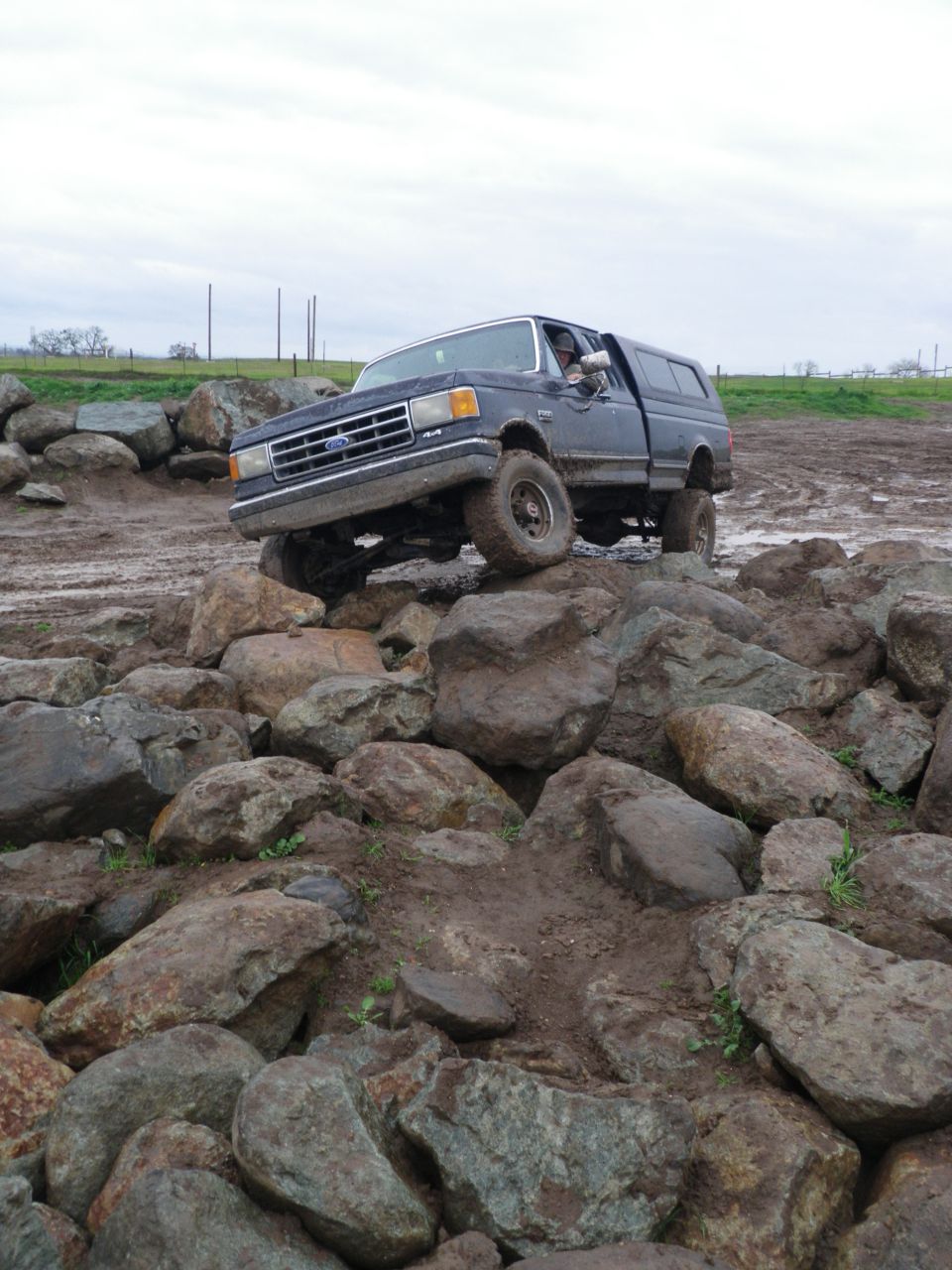 Dark blue Ford F-Series truck with a camper shell navigating a rocky, muddy terrain. The grille and headlights are visible