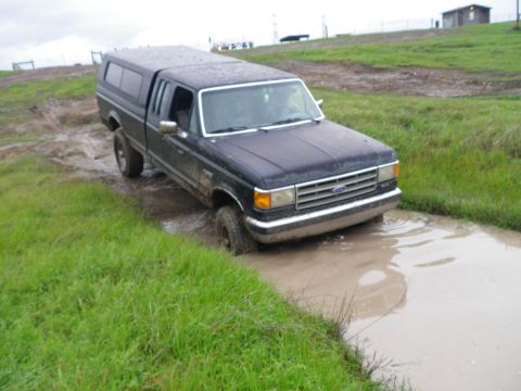1991 Ford Bronco - This dark blue pickup truck navigates a muddy path, its front end submerged in water.