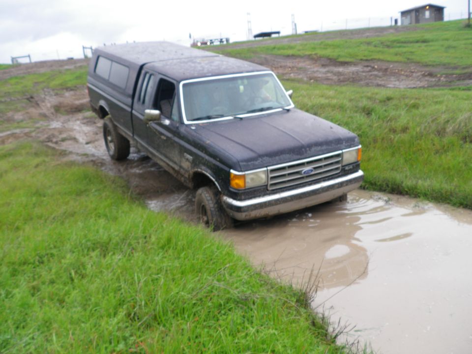 Dark blue pickup truck with camper shell stuck in mud, front 3/4 view. Chrome grille and bumper visible