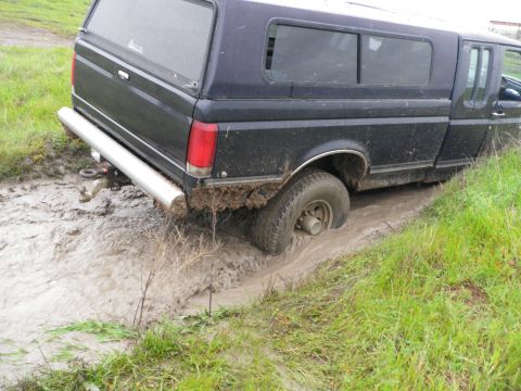 1991 Ford Bronco - A dark blue pickup truck with a camper shell is stuck in a muddy ditch, with its rear tire submerged.