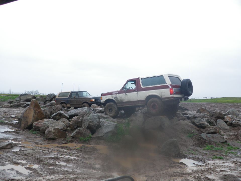 1991 Ford Bronco and another Bronco navigating a muddy obstacle course with large rocks