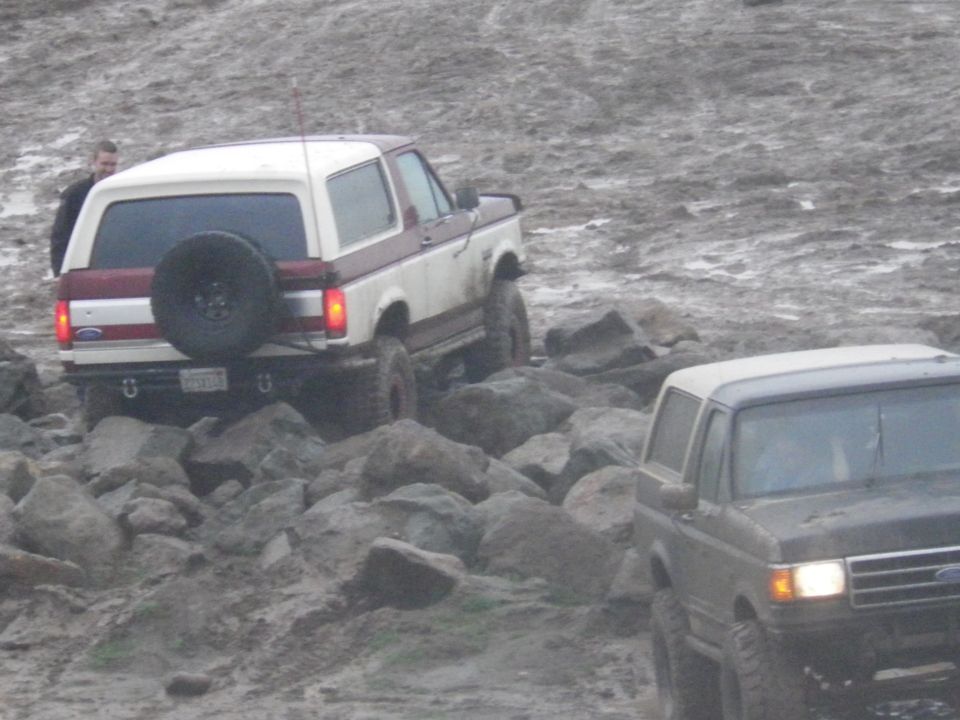 Two Ford Broncos navigating a rocky, muddy terrain. One is white and maroon, seen from the rear with a spare tire mounted. The other is dark, seen from the front-right