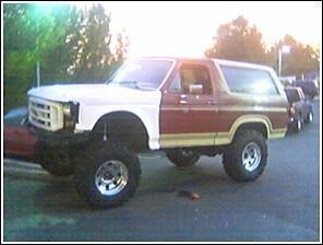 1986 Ford Bronco - This lifted 1980s Ford Bronco features a two-tone paint scheme and prominent off-road tires, with other vehicles visible in the background.