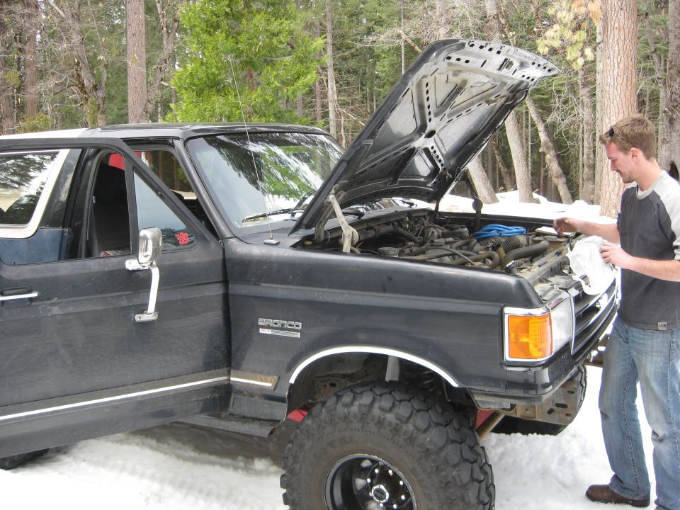 1986 Ford Bronco with hood open, man checking engine bay in snowy woods