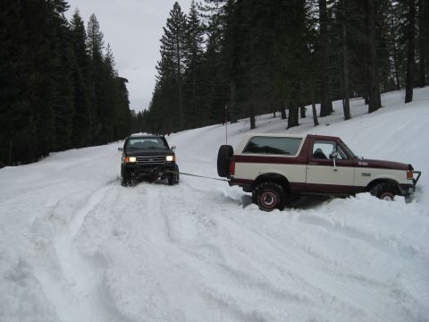 1986 Ford Bronco - A maroon and cream Ford Bronco is being towed by a black SUV through a snowy forest road.