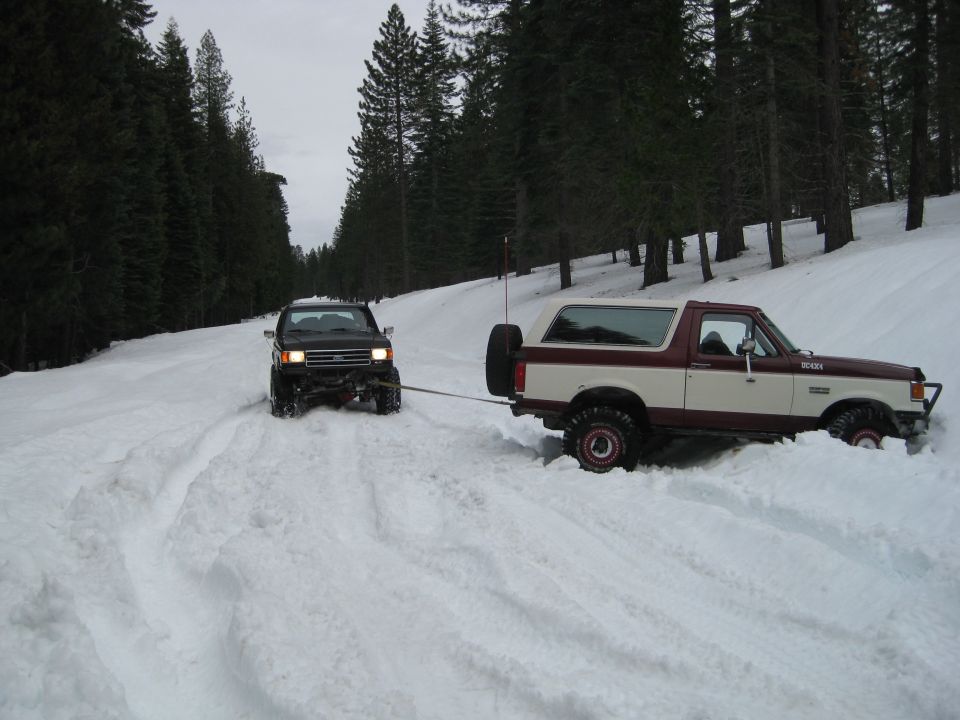 Two SUVs in snow, one maroon and cream Ford Bronco being towed by a black SUV