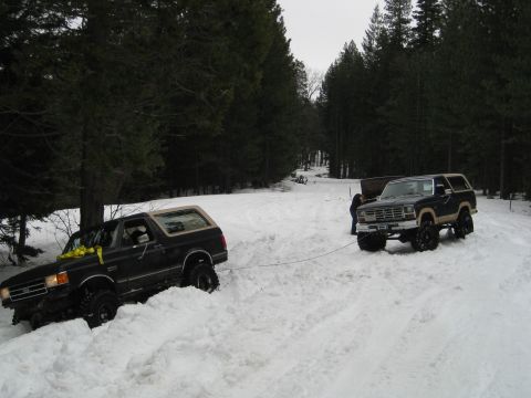 1986 Ford Bronco - Two Ford Broncos navigate a snowy trail, with one vehicle assisting the other out of a snowdrift.