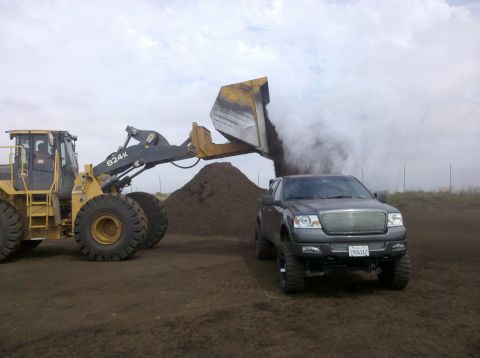 2005 Ford F-150 - A gray Ford F-150 truck is parked next to a front loader that is dumping a large pile of dirt.