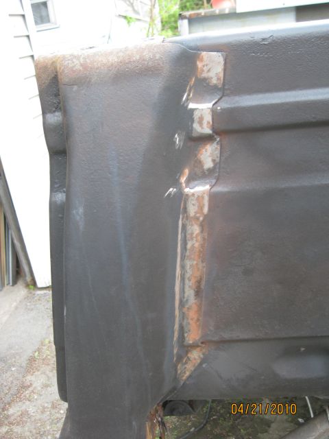 1983 Ford Bronco - Rear corner of a dark-painted body tub photographed against a white-sided house, showing the vertical seam where a quarter panel meets the…