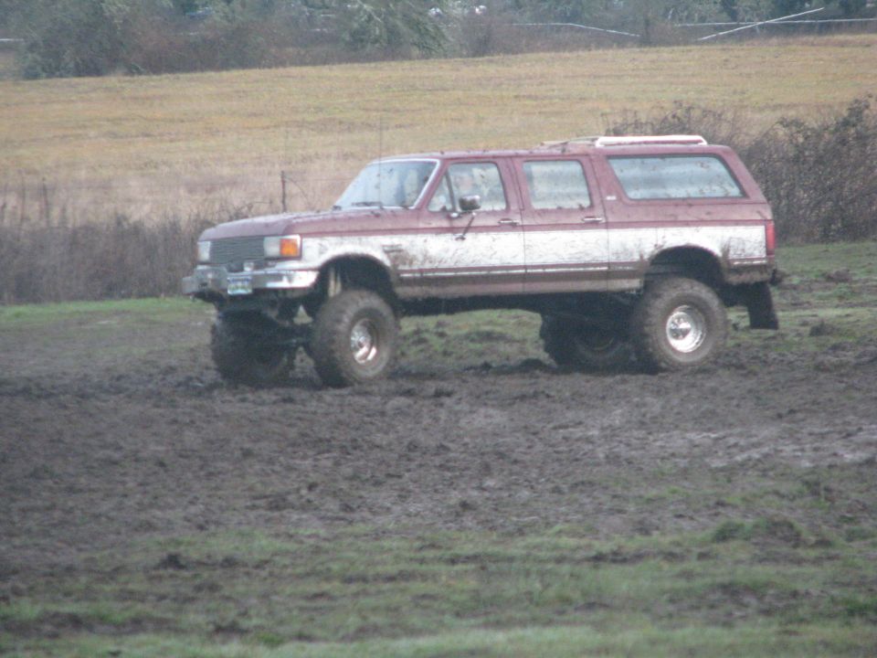 Lifted maroon and white four-door SUV with large tires driving through mud. Appears to be a 1990s Ford Bronco or similar