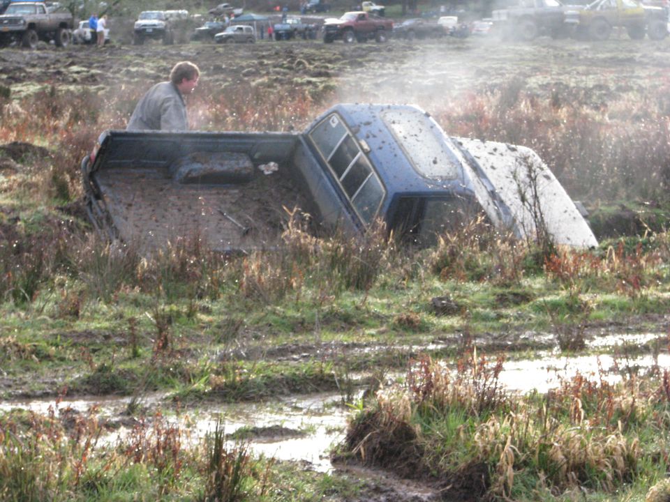 Blue pickup truck on its side in mud, with a person looking on