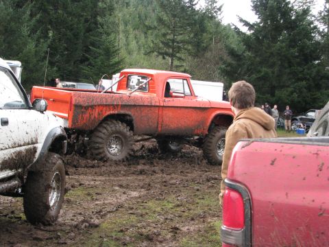 1978 Ford Bronco - An orange pickup truck with oversized tires is stuck in the mud, with other vehicles and people around.