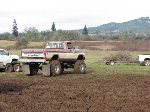 1978 Ford Bronco - A modified Ford pickup truck with oversized tires pulls a trailer across a muddy terrain under an overcast sky.