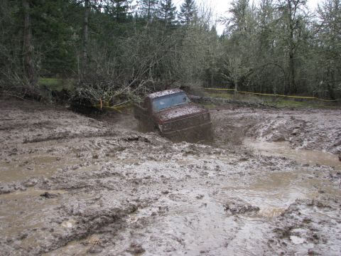 1978 Ford Bronco - A red pickup truck navigates a challenging muddy terrain, with trees and brush lining the background.