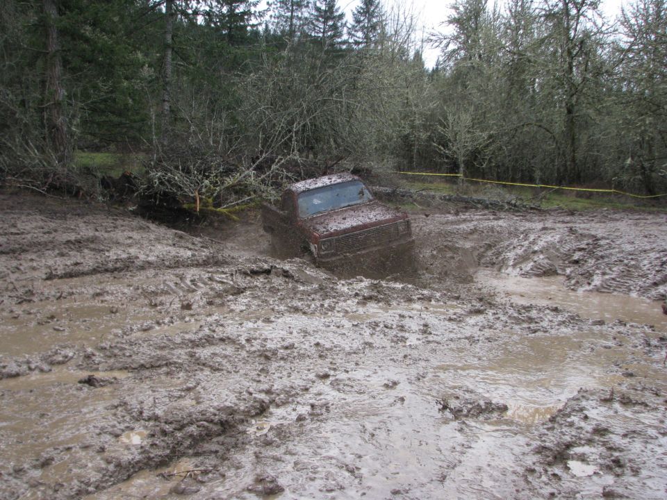 Red pickup truck struggling in deep mud, trees and brush in background