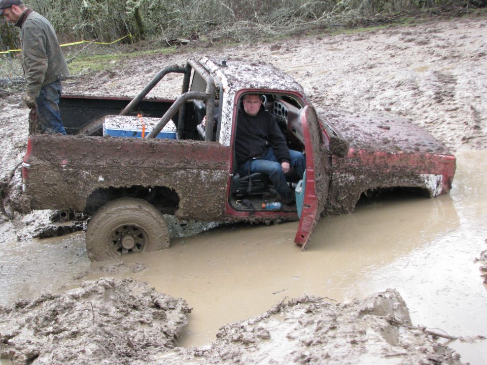Mud-covered red pickup truck stuck in mud, driver inside