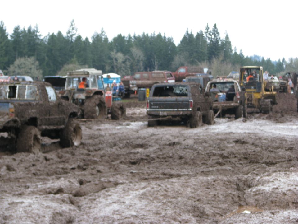 Muddy trucks and a yellow tractor in a muddy field with trees in the background