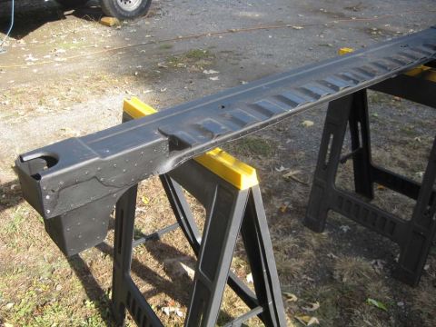 1983 Ford Bronco - A long refinished body support rail in dark gray primer laid across three yellow-and-black plastic sawhorses on a lawn, showing a boxed end…
