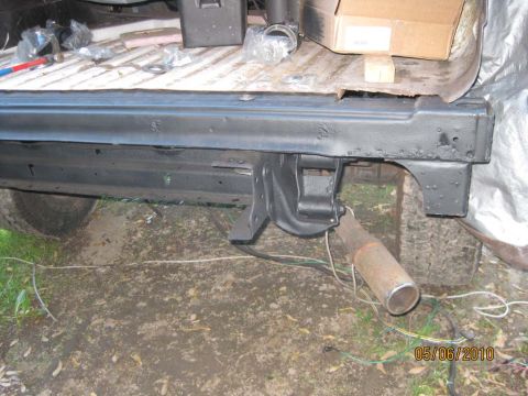 1983 Ford Bronco - Side view of a freshly black-painted body rail and rear wheel-arch section leaning against a stripped body tub outdoors, with a rusty exhau…