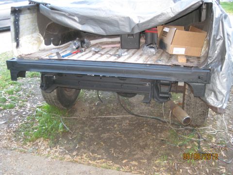 1983 Ford Bronco - Wide side view of a tarp-covered project vehicle on knobby tires with a long freshly black-painted body rail propped along its flank, a rus…