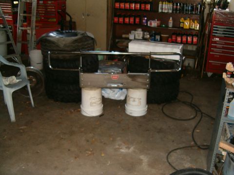 1991 Ford Bronco - A winch bumper is displayed in a garage, supported by buckets, with large tires stacked behind it.